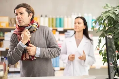 Mężczyzna w szaliku patrzy zmartwiony przy lady apteki, farmaceutka w białym fartuchu w tle.