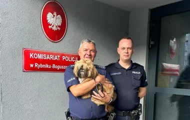 Dwaj policjanci w mundurach stoją przed komisariatem policji w Rybniku-Boguszowicach, jeden z nich trzyma na rękach małego psa.