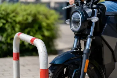 Przód czarnego motocykla stoi przy barierce ochronnej na brukowanej nawierzchni, w tle zielone krzewy.